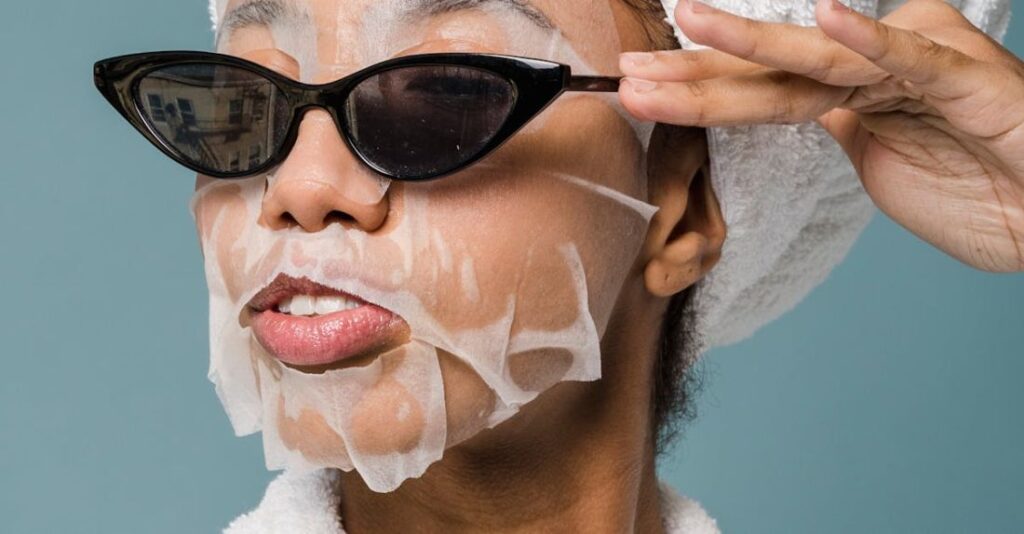 Hydrated - Self esteem young African American lady with moisturizing sheet mask and towel on hand wearing trendy sunglasses while standing against blue background in bathrobe after shower