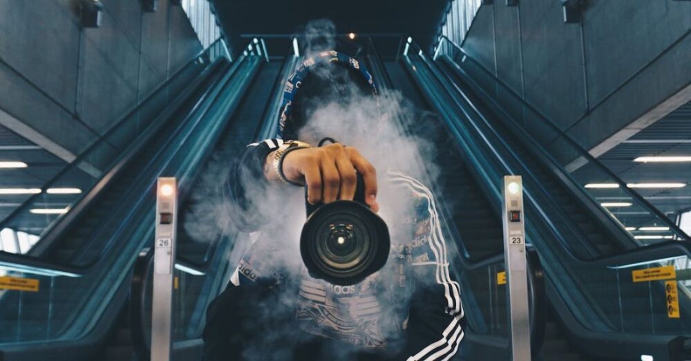 Wear - Man Holding Camera While Squating With Smoke on Face Inside Building